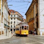 Image of one of Lisbon's iconic yellow tram cars driving through a cobblestone street line with yellow and white buildings.