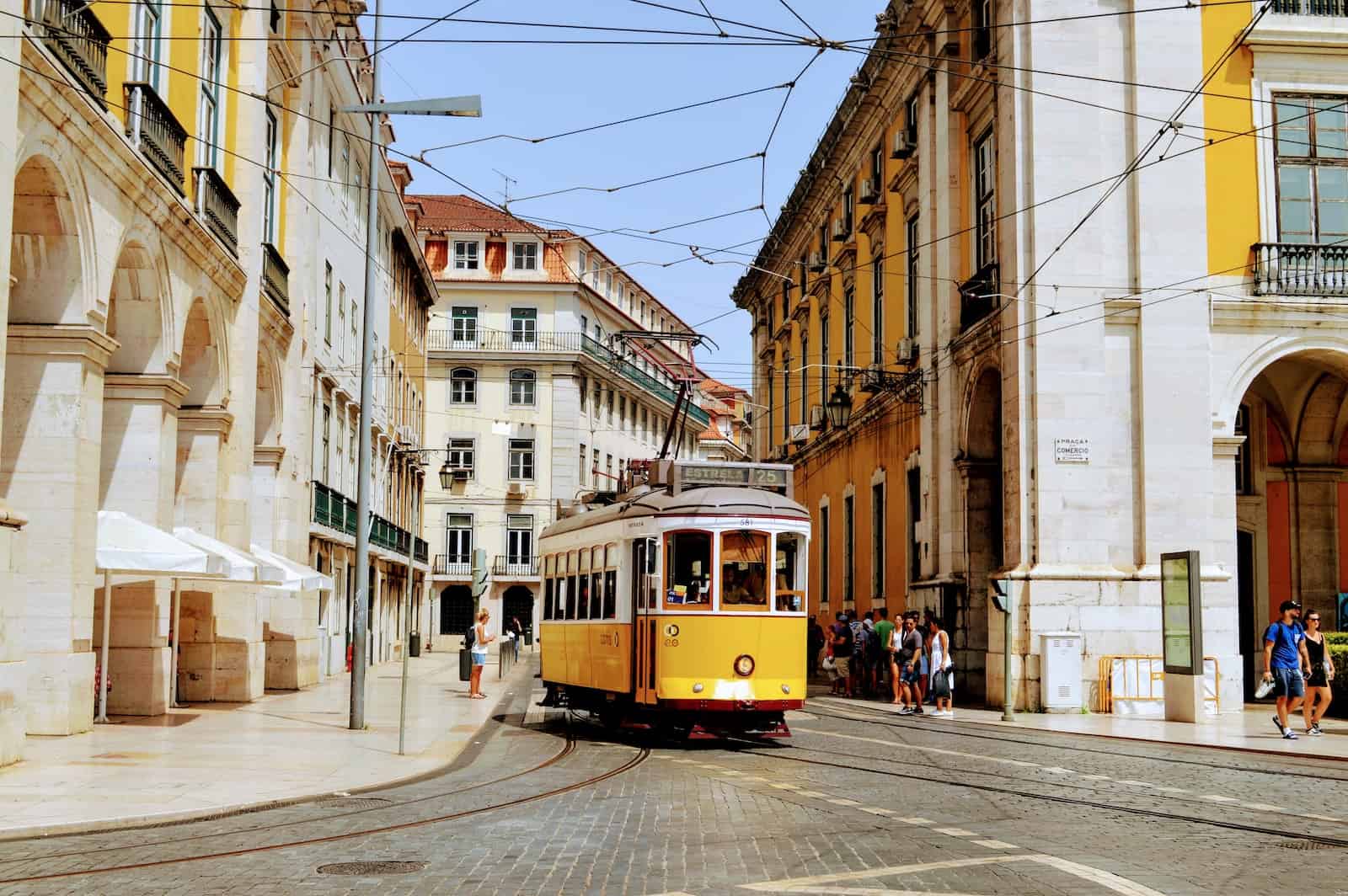 Image of one of Lisbon's iconic yellow tram cars driving through a cobblestone street line with yellow and white buildings.