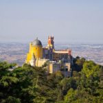 Pena palace in sintra, portugal, stands atop a hill surrounded by greenery, with the countryside stretching into the distance.