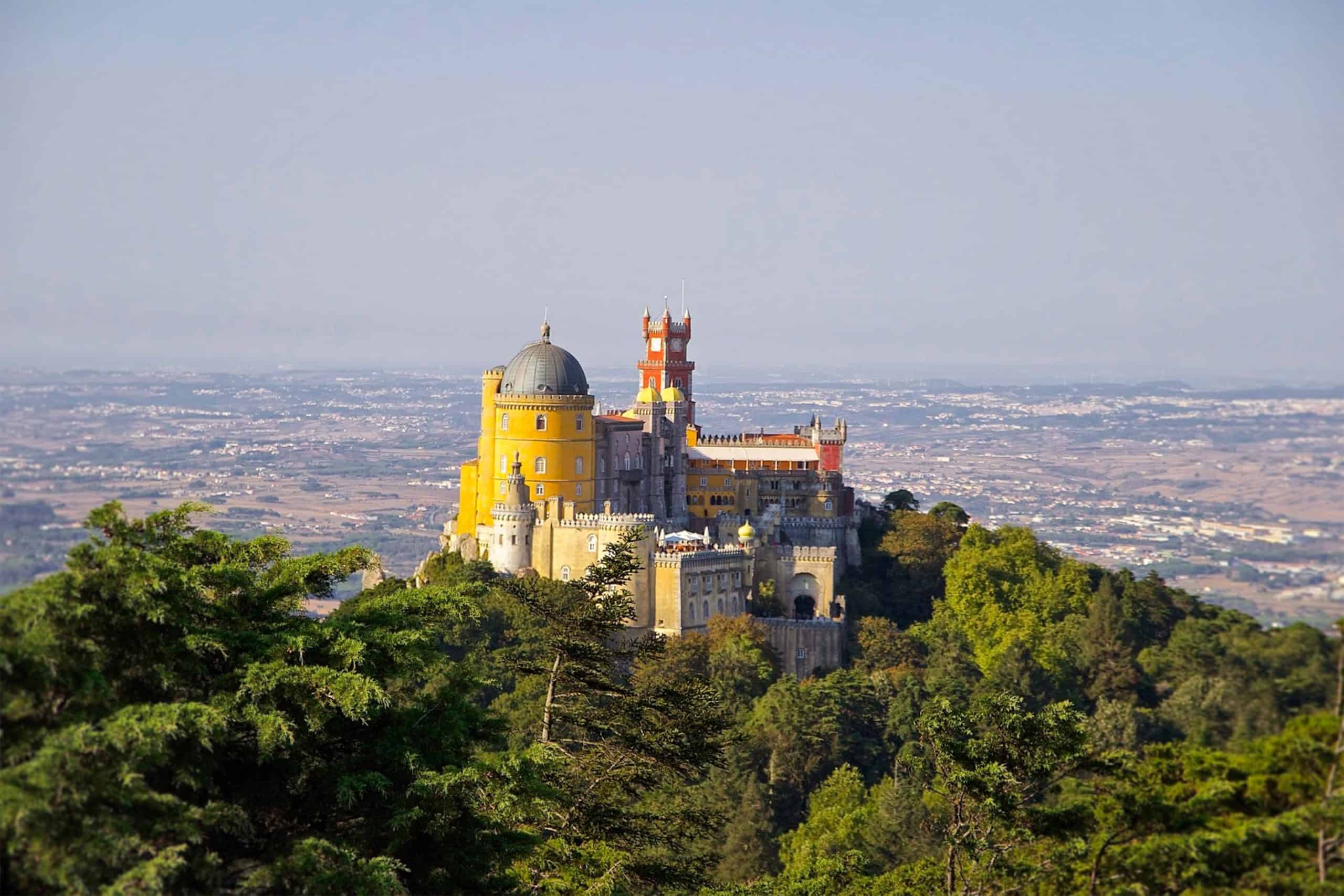 Pena palace in sintra, portugal, stands atop a hill surrounded by greenery, with the countryside stretching into the distance.