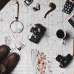 Travel and photography essentials laid out on a map, including a vintage camera, lenses, notebook, and a cup of coffee.