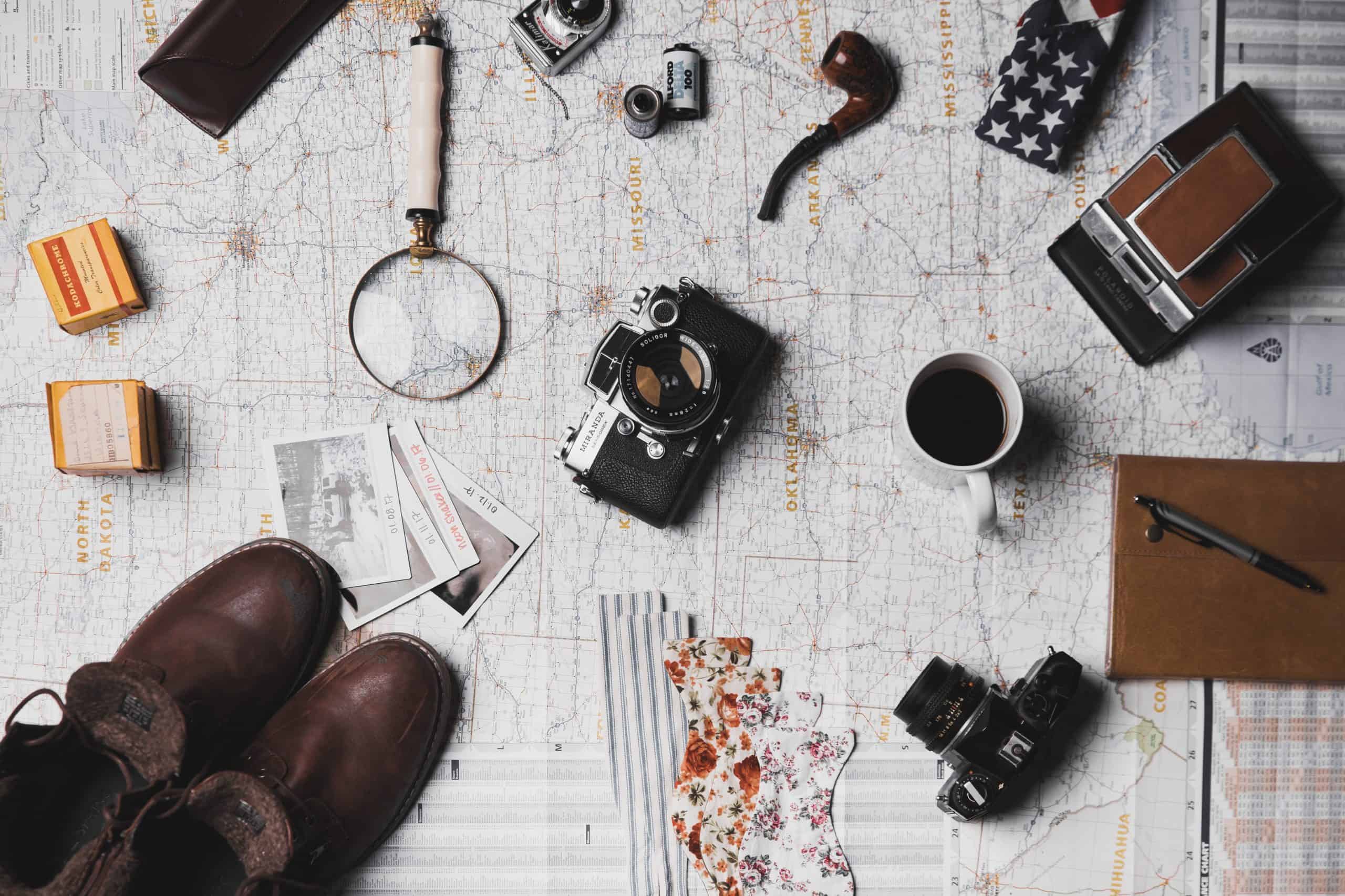 Travel and photography essentials laid out on a map, including a vintage camera, lenses, notebook, and a cup of coffee.