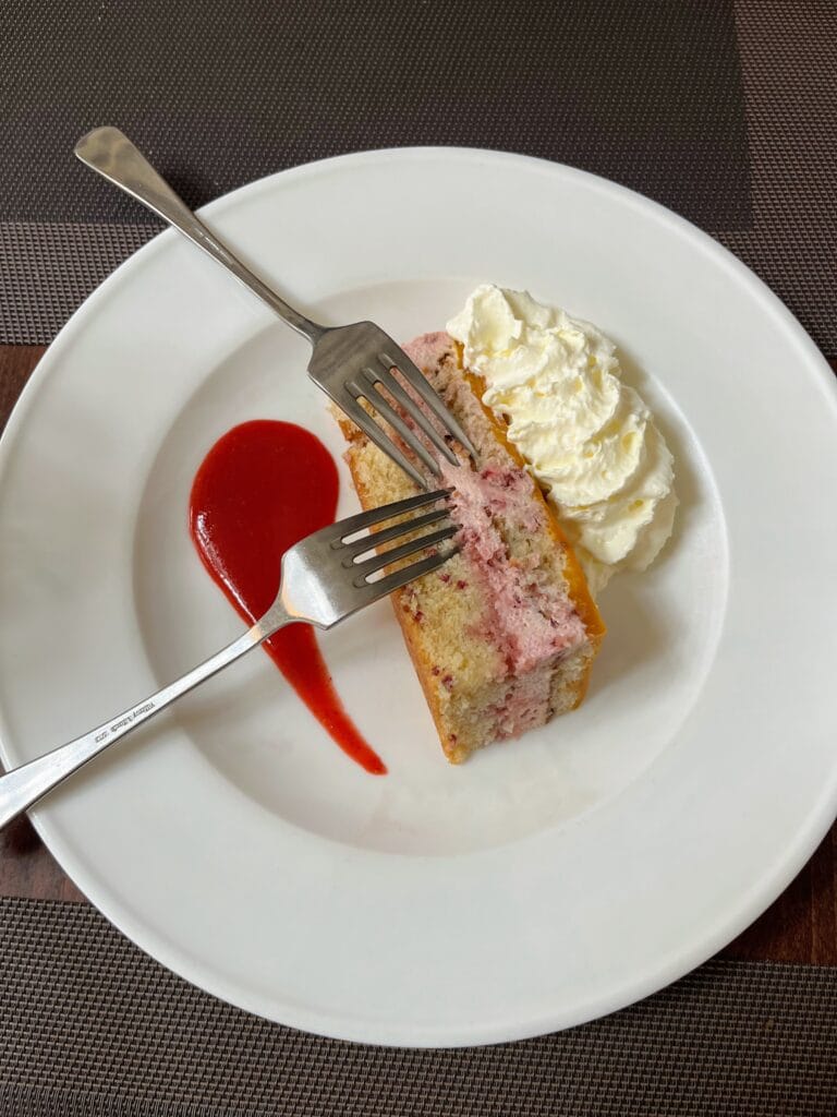 A slice of berry cake with whipped cream and strawberry sauce on a white plate, with two forks resting on top.