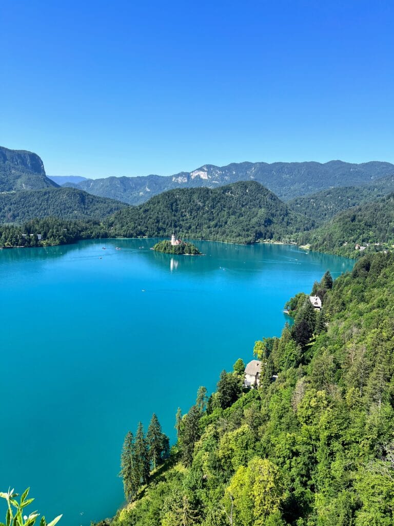 A tranquil turquoise lake surrounded by lush green forests with hills in the background on a clear day.