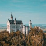 A fairytale castle perched atop a forest-covered hill under a clear blue sky.