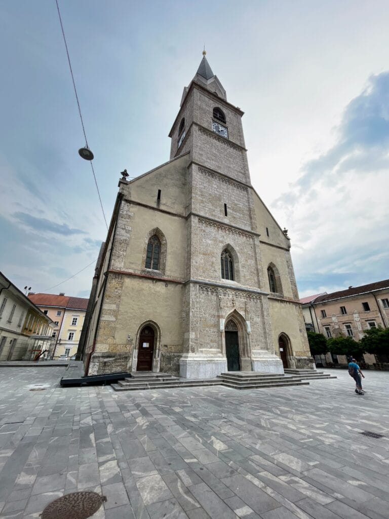 An old church with a prominent bell tower stands in a spacious city square under a cloudy sky.