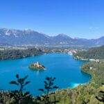 A panoramic view of lake bled with its island church, surrounded by lush greenery and a backdrop of mountain ranges under a clear blue sky.