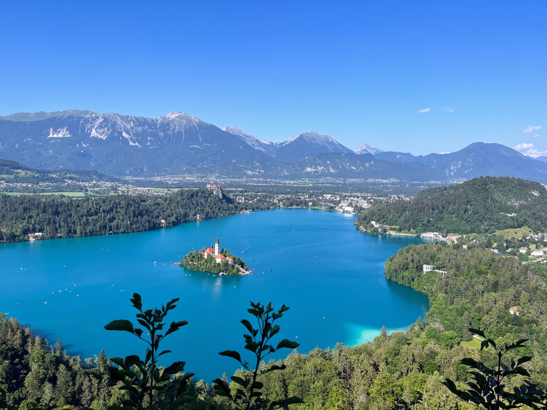 A panoramic view of lake bled with its island church, surrounded by lush greenery and a backdrop of mountain ranges under a clear blue sky.