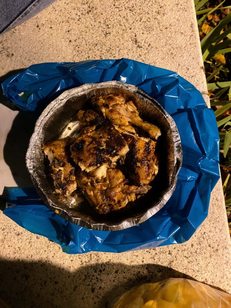 Grilled chicken pieces served hot in a disposable aluminum tray on an outdoor surface, with a blue plastic bag
