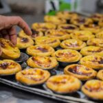 A person selecting a freshly baked pastel de nata from a tray full of portuguese custard tarts.