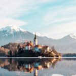 A serene lake reflecting a church with a backdrop of mountains under a clear sky.