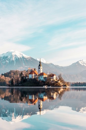 A serene lake reflecting a church with a backdrop of mountains under a clear sky.