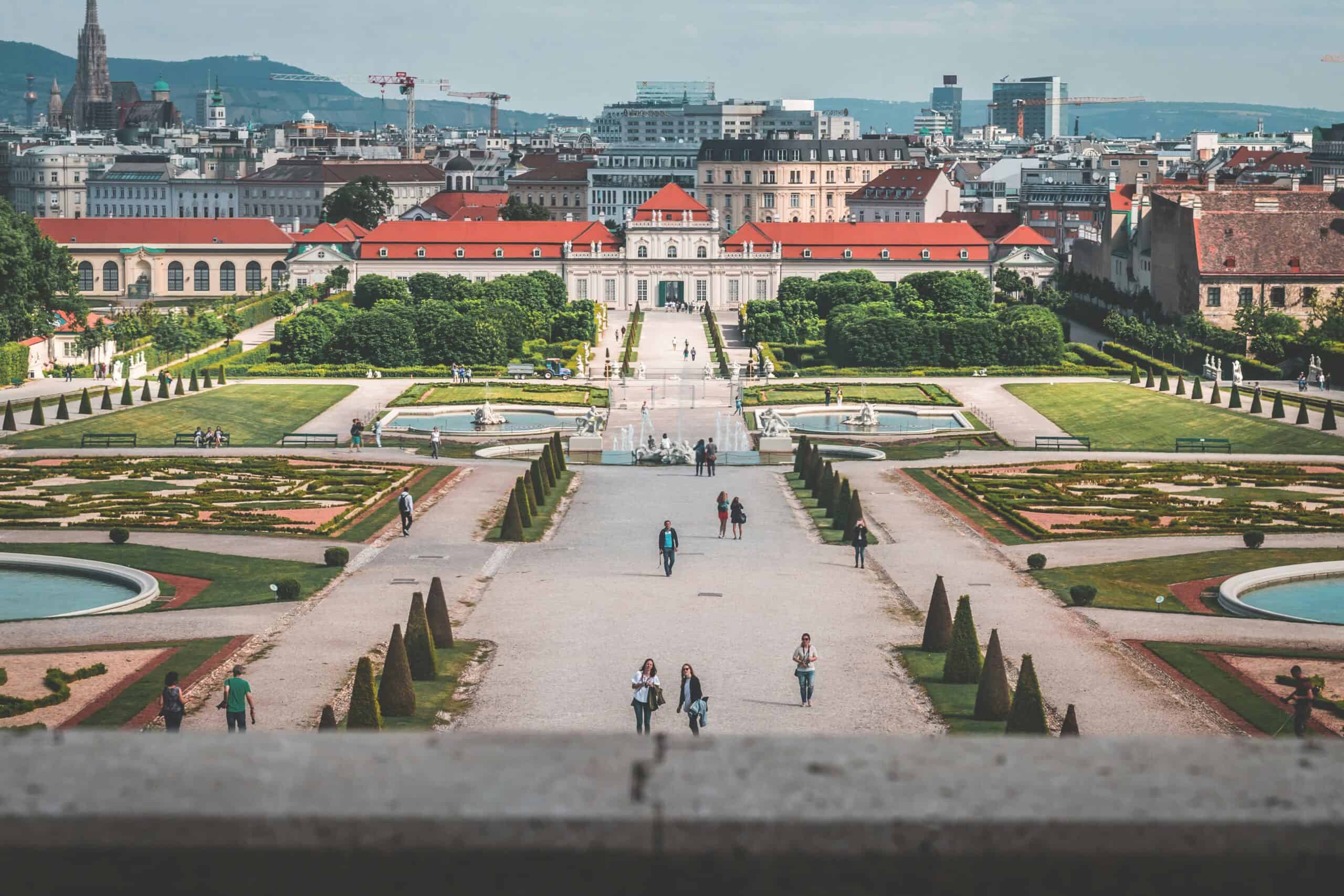 Formal gardens and walking paths leading to a classical palace with the city skyline in the background.