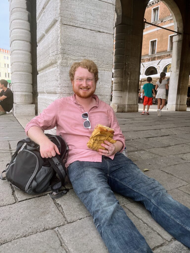 A man in a red button down and jeans sitting on a cobblestone street against a stone pillar holding a sandwich