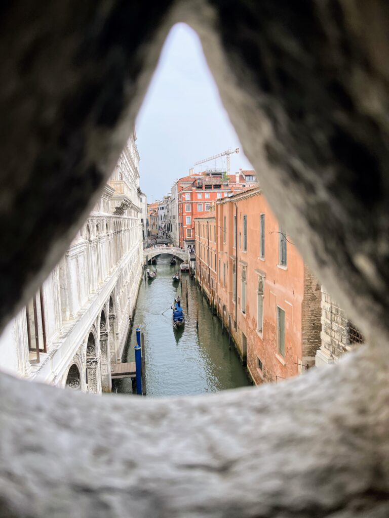 A view through a triangular hole over a canal with historic buildings, boats, and a bridge in the background