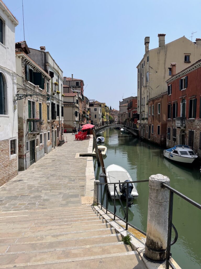 A stone street next to a canal with historic buildings on either side and a wooden bridge in the distance