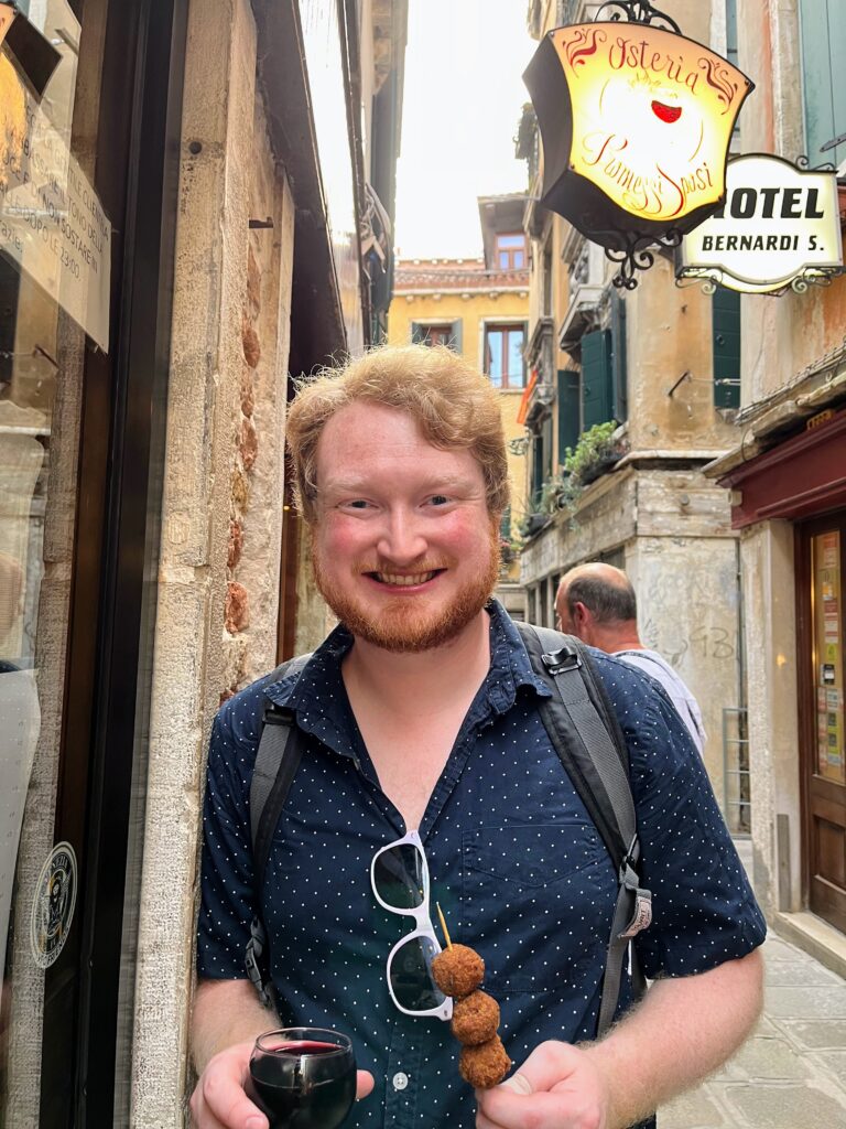 A man standing in an Italian street holding a glass of red wine and a skewer with meatballs