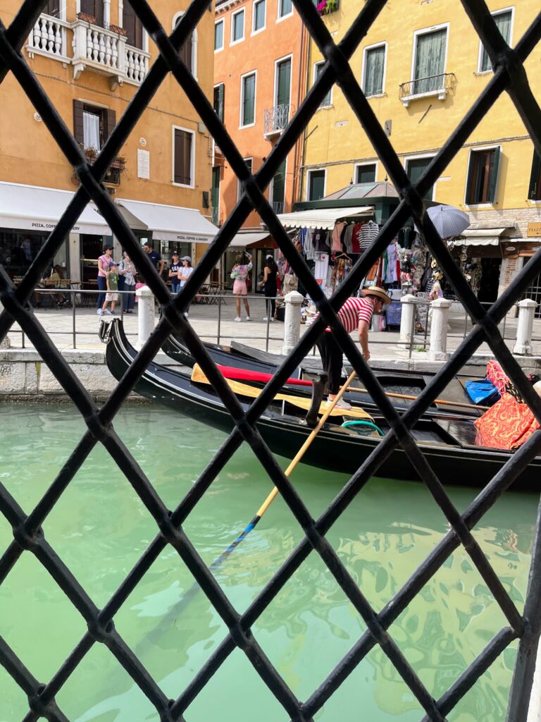 A view through a caged window onto a Venetian canal with a gondolier steering a boat