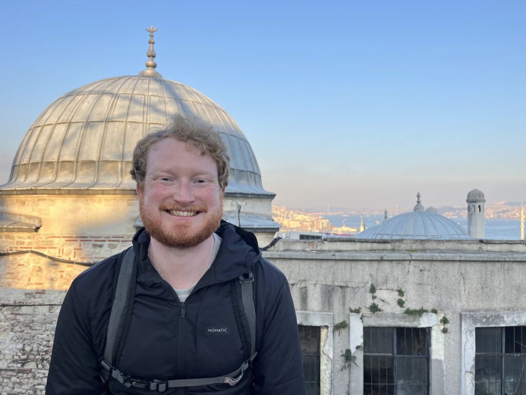 A white man with short wavy red hair and a beard, wearing a black jacket. In the background is a mosque and a city skyline along a river.