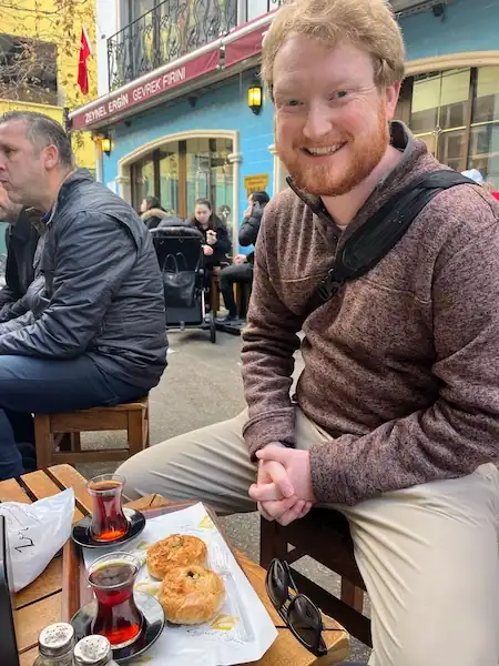 Man sitting at a small wooden table with a tray that has two cups of black tea and two round pastries.